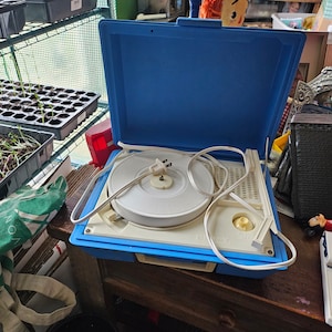 May include: A blue and white portable record player with a white turntable and a cord with a plug. The record player is open and the lid is propped up behind it.