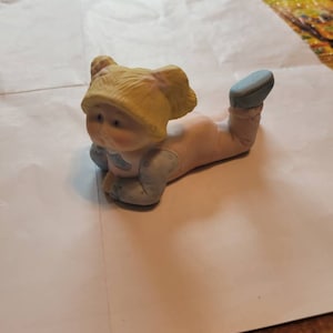 May include: A ceramic figurine of a Cabbage Patch Kid doll lying on its stomach. The doll has blonde hair, a pink dress, and blue shoes.