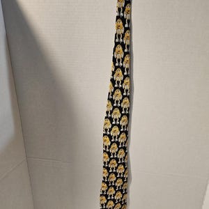 Può includere: Una cravatta nera con un motivo ripetuto di personaggi dei cartoni animati gialli. I personaggi hanno facce, braccia e gambe bianche, con corpi gialli. La cravatta è realizzata in tessuto liscio ed è adatta per occasioni formali o informali.