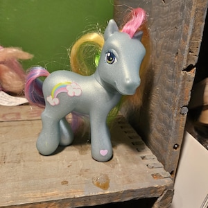 Puede incluir: Un My Little Pony azul claro con una crin y cola rosa, amarilla y azul. El pony tiene un símbolo de arcoíris y nubes en su flanco y un corazón rosa en su pezuña.
