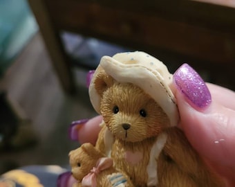 May include: A brown teddy bear wearing a white hat with blue polka dots is holding a smaller brown teddy bear. The larger bear is wearing a pink heart and a white bib.