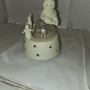 Puede incluir: Una figura musical blanca con una figura sentada con un traje de nieve sobre una cúpula de cristal. Hay dos pequeñas figuras de pingüinos blancos. La base es blanca con recortes de estrellas.