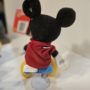 May include: A Mickey Mouse plush doll, seen from the back, with a red jacket, blue shorts, and yellow shoes. The doll has black ears and white gloves. The doll is mounted on a grey stand. The tag reads "Surface Washable Only".