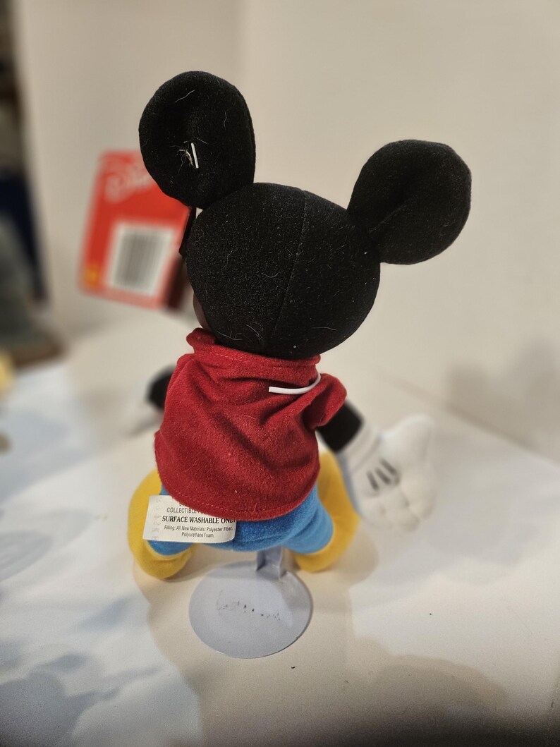 May include: A Mickey Mouse plush doll, seen from the back, with a red jacket, blue shorts, and yellow shoes. The doll has black ears and white gloves. The doll is mounted on a grey stand. The tag reads "Surface Washable Only".