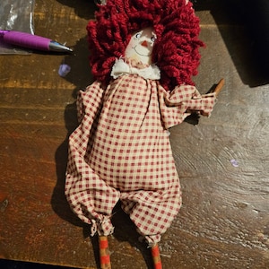 Könnte beinhalten: Eine handgemachte Stoffpuppe mit roten Wollhaaren, einem rot-weiß karierten Kleid und braunen Holzbeinen.