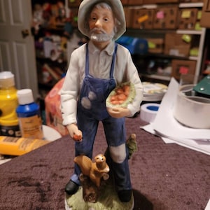 Könnte beinhalten: Keramikfigur eines älteren Mannes in blauen Latzhosen und einem weißen Hemd, der eine Wassermelone und einen Apfel hält. Ein Eichhörnchen sitzt auf einem Baumstumpf zu seinen Füßen. Die Figur hat ein verwittertes Aussehen.