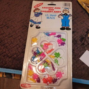 May include: A plastic package containing a set of colorful snap beads featuring Raggedy Ann and Andy. The beads are shaped like hearts, stars, and other fun shapes. The package includes the text "Raggedy Ann and Raggedy Andy Li'l Snap Beads".