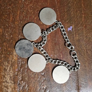 May include: A silver-toned charm bracelet featuring five circular charms connected by a chain. The charms have a brushed metal appearance, with one showing signs of wear. The bracelet is displayed on a wooden surface.