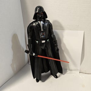 以下が含まれることがあります： 赤いライトセーバーを持った黒いダース・ベイダーのアクションフィギュア。フィギュアは黒いヘルメット、胸当て、マントを着用しています。ライトセーバーは赤く、フィギュアの右手で持っています。フィギュアは直立しています。