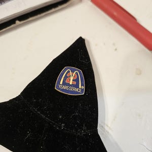 Puede incluir: Un pin con el logotipo de McDonald's y el texto "2 Years Service" sobre un fondo azul. El pin está sobre un fondo de terciopelo negro. Un bolígrafo rojo y una pila de papeles están en el fondo.