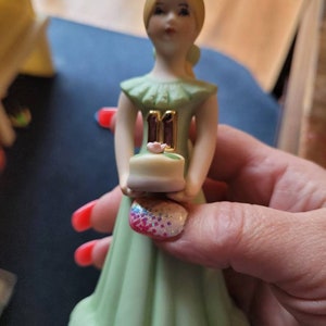 May include: A porcelain figurine of a girl in a green dress holding a small cake with the number "11" on top. The cake is decorated with a pink flower.