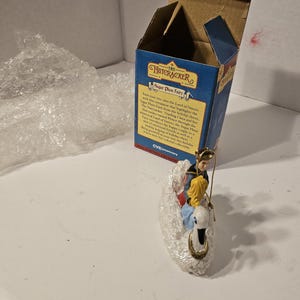 Op de afbeelding: Een witte porseleinen ornament met een scène uit De Notenkraker. Een prins en een jong meisje rijden op een witte zwaan. De ornament is verpakt in een blauwe doos met de woorden "The Nutcracker Sugar Plum Fairy" erop gedrukt.