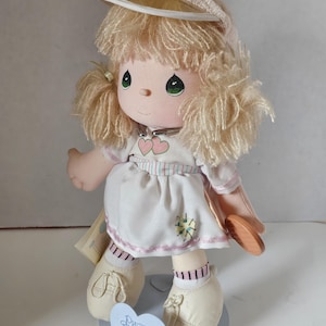 May include: A Precious Moments doll wearing a white dress with pink accents, a yellow visor, and white shoes. The doll is holding a tennis racket and has blonde hair. The doll is standing on a base with the words "Precious Moments Last Forever by Enesco" printed on it.