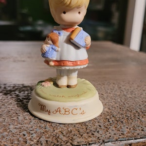 Może przedstawiać: Porcelanowa figurka dziewczynki z blond włosami, która nosi czerwoną kokardkę, niebiesko-białą sukienkę i niebieską książkę. Trzyma lalkę w lewej ręce. Figurka stoi na białej podstawie z napisem "My ABC's" i "You watch and you'll learn".
