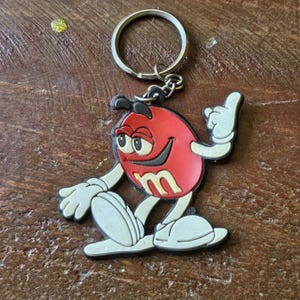 Puede incluir: Llavero de personaje M&M rojo con una sonrisa blanca y un contorno negro. El llavero está hecho de goma y tiene un anillo de llave plateado.