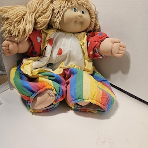 Op de afbeelding: Een vintage pop met blond garen haar, een geschilderd gezicht en een clownachtig outfit. De outfit van de pop bevat een witte top met rode stippen, een gele gerimpelde kraag, rode mouwen en een regenboog gestreepte broek.