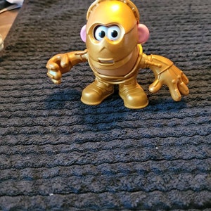 Pode incluir: Uma figura de brinquedo de plástico dourado de um robô com cabeça de batata. O robô tem uma cabeça redonda com um rosto, olhos grandes e uma boca pequena. O robô tem dois braços e duas pernas. O robô está em pé sobre uma superfície preta.