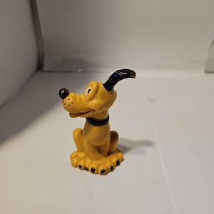 Könnte beinhalten: Eine gelbe Figur von Pluto, einem Disney-Zeichentrickhund, in sitzender Position. Die Figur hat schwarze Ohren, eine schwarze Nase und einen schwarzen Kragen. Der Hund sitzt auf einem Sockel mit schwarzen Pfoten.