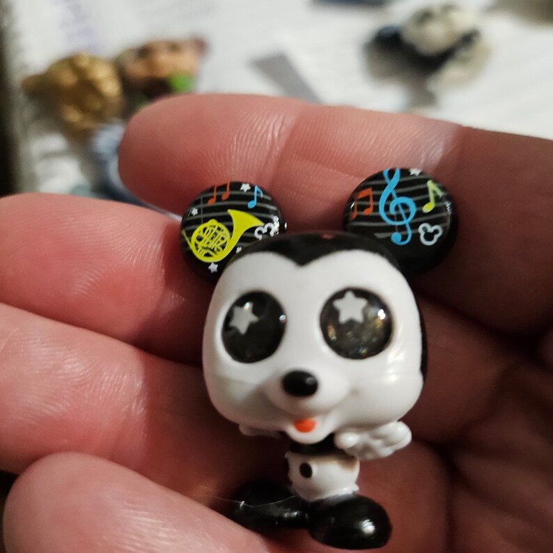 Peut inclure: Une figurine de Mickey Mouse noire et blanche avec de grands yeux blancs orn&eacute;s d'&eacute;toiles argent&eacute;es. Mickey porte des chaussures noires et a une bouche rouge. Ses oreilles sont noires avec des notes de musique et une trompette jaune sur une oreille.