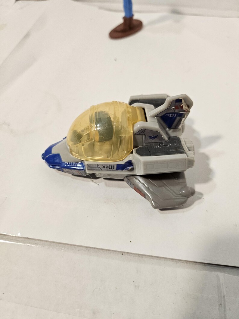 May include: A toy spaceship in shades of gray, blue, and white, with a transparent yellow cockpit. The toy has the text "XL-01" printed on the side. The spaceship is a miniature replica of a fictional vehicle.