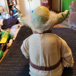 K&ouml;nnte beinhalten: Eine gr&uuml;ne Yoda-Figur mit wei&szlig;em Bart und braunem Gewand. Die Figur tr&auml;gt einen braunen G&uuml;rtel und hat einen Fransenbesatz.