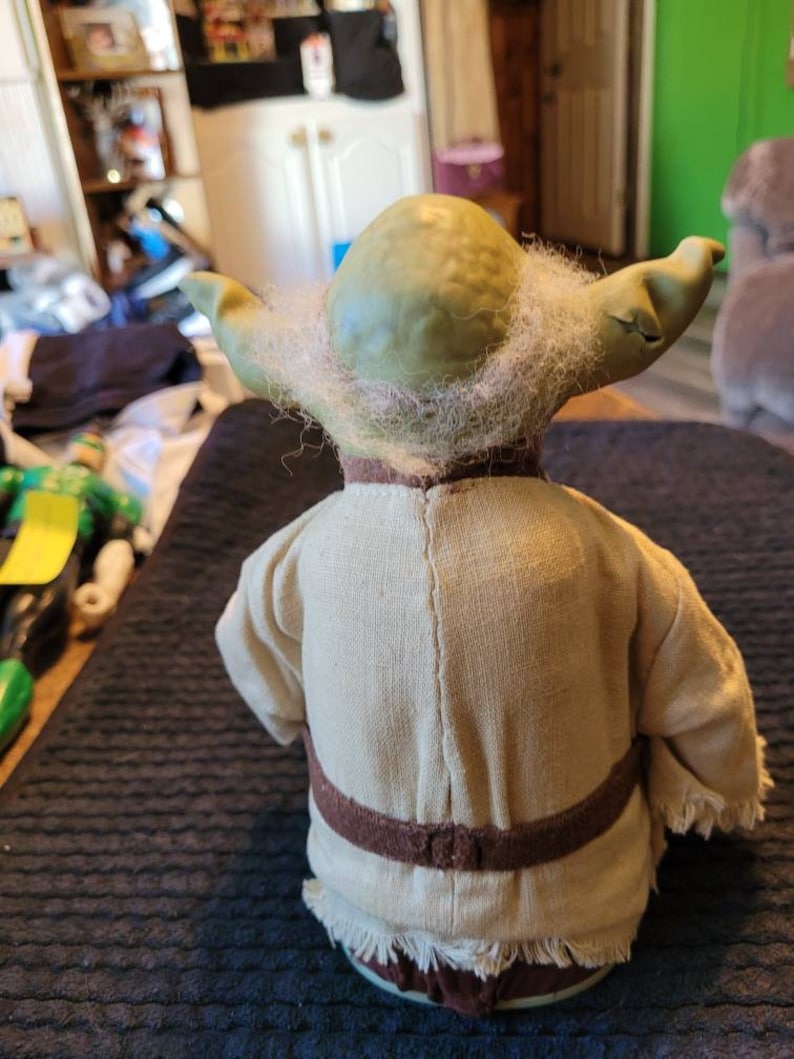 K&ouml;nnte beinhalten: Eine gr&uuml;ne Yoda-Figur mit wei&szlig;em Bart und braunem Gewand. Die Figur tr&auml;gt einen braunen G&uuml;rtel und hat einen Fransenbesatz.