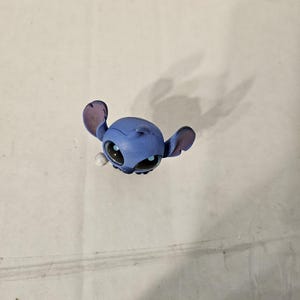 Pu&ograve; includere: Una piccola statuina blu del personaggio Stitch del film d'animazione Lilo & Stitch. La statuina ha grandi orecchie, occhi neri e un'espressione facciale dettagliata. La statuina &egrave; su uno sfondo semplice.