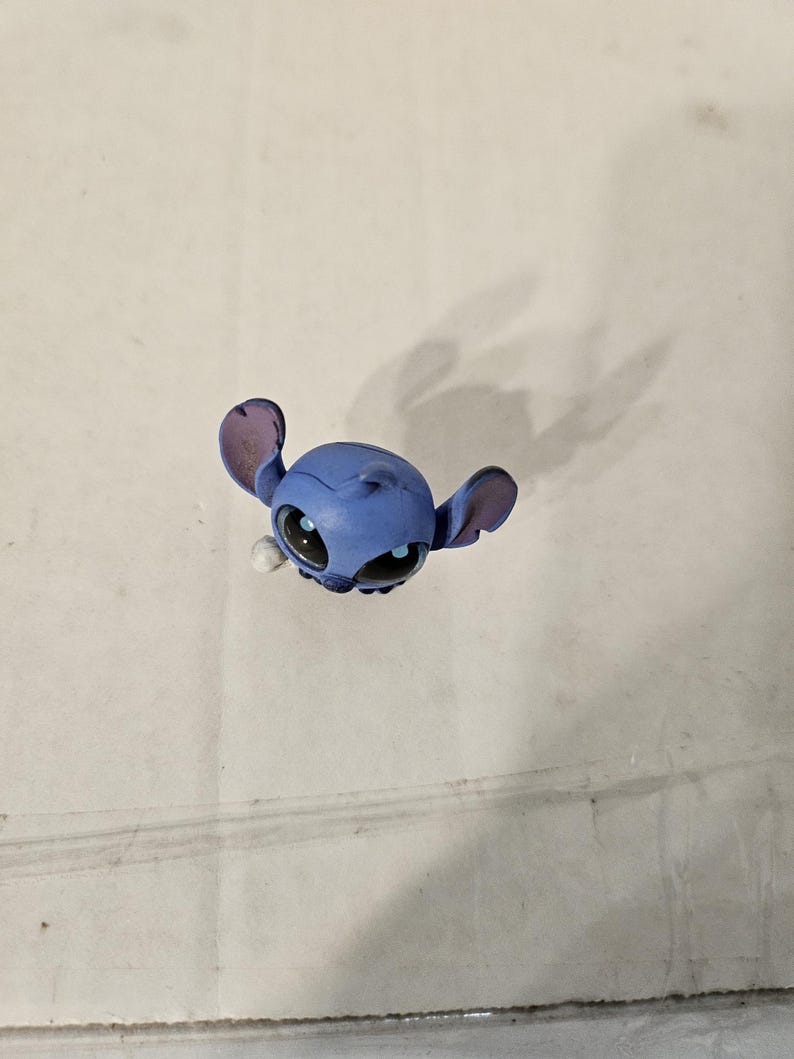 Pu&ograve; includere: Una piccola statuina blu del personaggio Stitch del film d'animazione Lilo & Stitch. La statuina ha grandi orecchie, occhi neri e un'espressione facciale dettagliata. La statuina &egrave; su uno sfondo semplice.