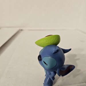 May include: A blue Stitch figurine from Lilo & Stitch, with a green avocado slice balanced on its head. The figurine is in a playful pose, with its arms and legs slightly bent. The avocado has a brown pit.