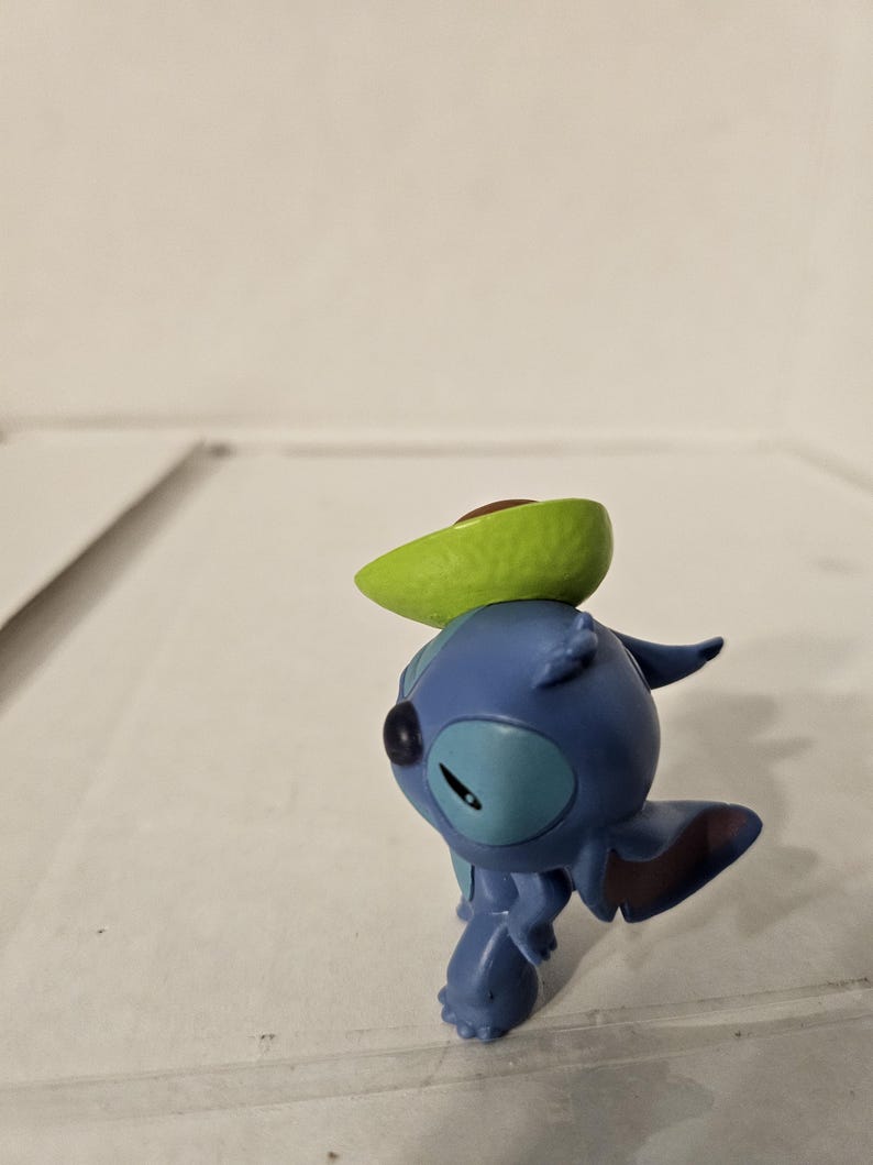 May include: A blue Stitch figurine from Lilo & Stitch, with a green avocado slice balanced on its head. The figurine is in a playful pose, with its arms and legs slightly bent. The avocado has a brown pit.