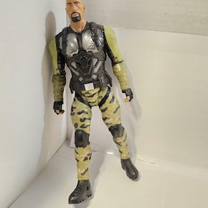 May include: Action figure of a bald man with a goatee, wearing a military-style outfit. The figure has a black and silver chest piece, olive green sleeves, and camouflage pants. Black boots and gloves complete the look. The figure is posed in a dynamic stance.