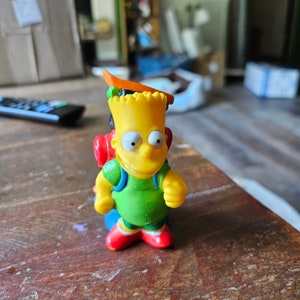 May include: A yellow plastic toy figure of a cartoon character wearing a green shirt and a red backpack. The character has a yellow face with black eyes and a wide smile.