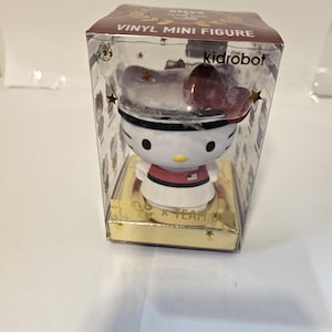 May include: A Kidrobot Vinyl Mini Figure of Hello Kitty wearing a white hat with a red bow and a red and white outfit. The figure is in a clear plastic box with a gold base and a brown top that reads "VINYL MINI FIGURE".