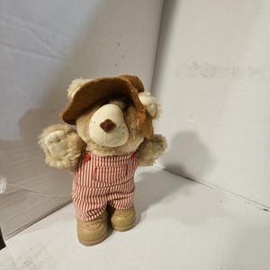 以下が含まれることがあります： 茶色の帽子と赤と白のストライプのオーバーオールを着たテディベアのぬいぐるみ。クマは薄茶色の鼻とタン色の靴をしています。クマの両腕は広げられており、直立しています。