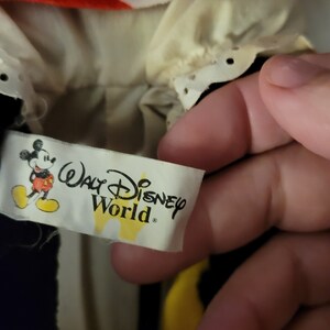 Puede incluir: Una etiqueta amarilla y blanca con el texto "Walt Disney World" y una imagen de dibujos animados de Mickey Mouse. La etiqueta est&aacute; unida a un juguete de peluche negro y rojo.