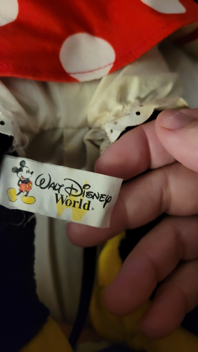 Puede incluir: Una etiqueta amarilla y blanca con el texto "Walt Disney World" y una imagen de dibujos animados de Mickey Mouse. La etiqueta est&aacute; unida a un juguete de peluche negro y rojo.