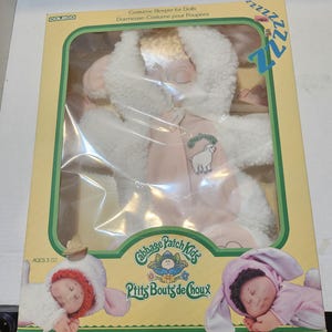 May include: A Cabbage Patch Kids doll costume sleeper in a box. The costume is white and pink, with a sheep graphic. The box has the text "Costume Sleeper for Dolls" and "P'tits Bouts de Choux". Ages 3+.