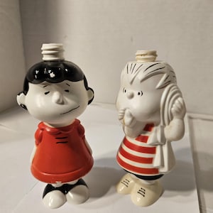 Puede incluir: Dos figuras de cerámica de personajes de dibujos animados. Una figura viste un vestido rojo, zapatos negros y tiene el pelo negro. La otra figura viste una camisa a rayas rojas y blancas, zapatos blancos y tiene el pelo blanco. Ambas figuras tienen una parte superior blanca.