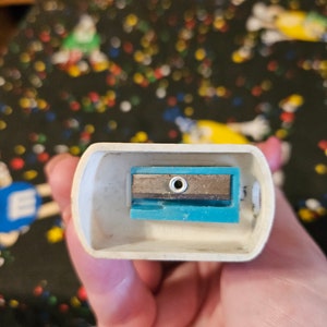 May include: A white plastic pencil sharpener with a blue and silver blade. The sharpener has a small hole in the center of the blade.