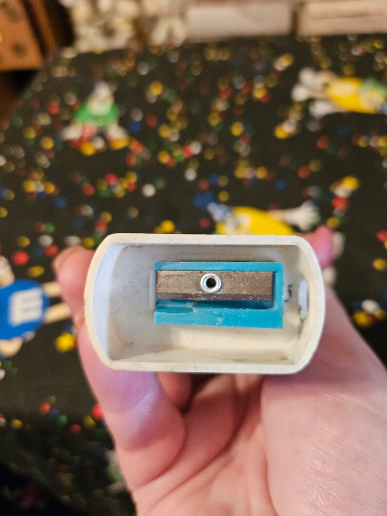 May include: A white plastic pencil sharpener with a blue and silver blade. The sharpener has a small hole in the center of the blade.