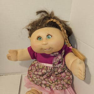 Puede incluir: Una muñeca vintage con cara redonda, ojos verdes y cabello castaño peinado en una trenza. La muñeca lleva una blusa magenta, una falda con estampado floral y una enagua de tul rosa. Los brazos de la muñeca están extendidos.