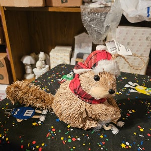 Könnte beinhalten: Ein brauner und weißer Eichhörnchen-Weihnachtsbaumschmuck mit einem rot-schwarzen karierten Schal und einer roten Weihnachtsmannmütze. Der Schmuck liegt auf einem schwarzen Hintergrund mit bunten Konfetti.