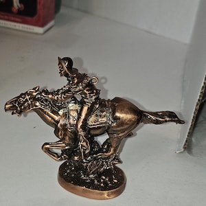 May include: A bronze-colored figurine of a rider on horseback. The rider wears a hat and detailed clothing, and the horse is in mid-gallop. The base is oval-shaped, and the overall design suggests a historical or decorative theme.