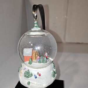 Peut inclure: Un globe de neige en verre transparent avec une scène d'hiver à l'intérieur. La base est blanche avec des arbres verts et des figurines. À l'intérieur, un bâtiment rouge et des figurines sont visibles. Un ruban noir est attaché au sommet.
