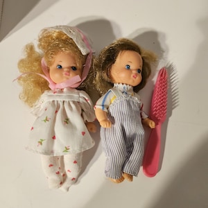 May include: Two small dolls with blonde and brown hair wearing clothes. The doll with blonde hair is wearing a white dress with pink flowers and a pink hat. The doll with brown hair is wearing blue overalls with white stripes. A pink plastic hairbrush is included.