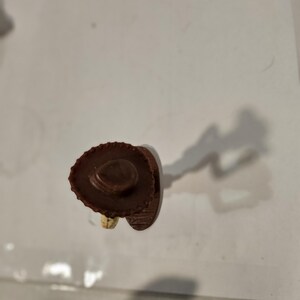 May include: A small, brown cowboy hat figurine with a wide brim and a detailed crown. The hat is made of a textured material, and the figurine is set against a plain background. The figurine is a miniature representation of a cowboy hat.