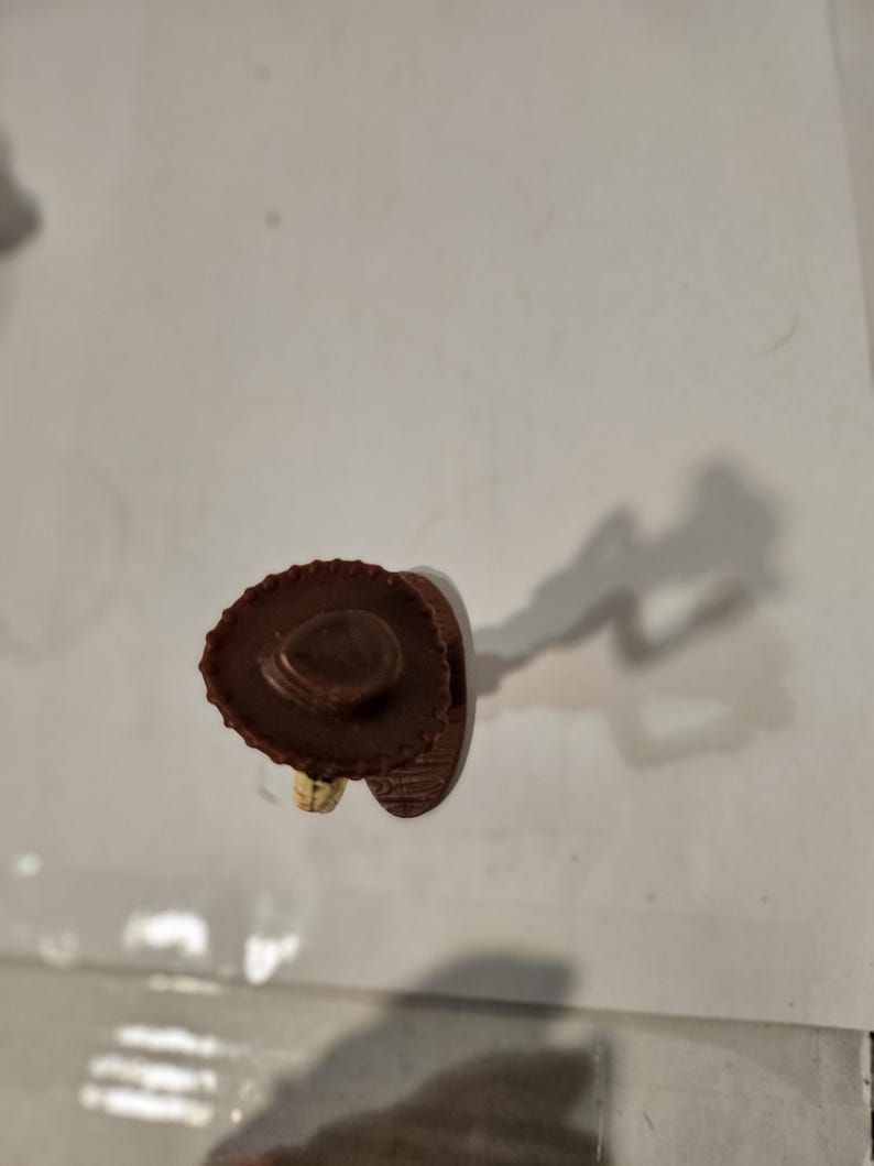 May include: A small, brown cowboy hat figurine with a wide brim and a detailed crown. The hat is made of a textured material, and the figurine is set against a plain background. The figurine is a miniature representation of a cowboy hat.