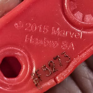 Może przedstawiać: Zbliżenie czerwonej plastikowej części zabawki z napisem "&copy; 2015 Marvel Hasbro SA" i numerem "#F3273" wygrawerowanym na powierzchni. Część ma zaokrąglony prostokątny kształt z okrągłymi i sześciokątnymi otworami.