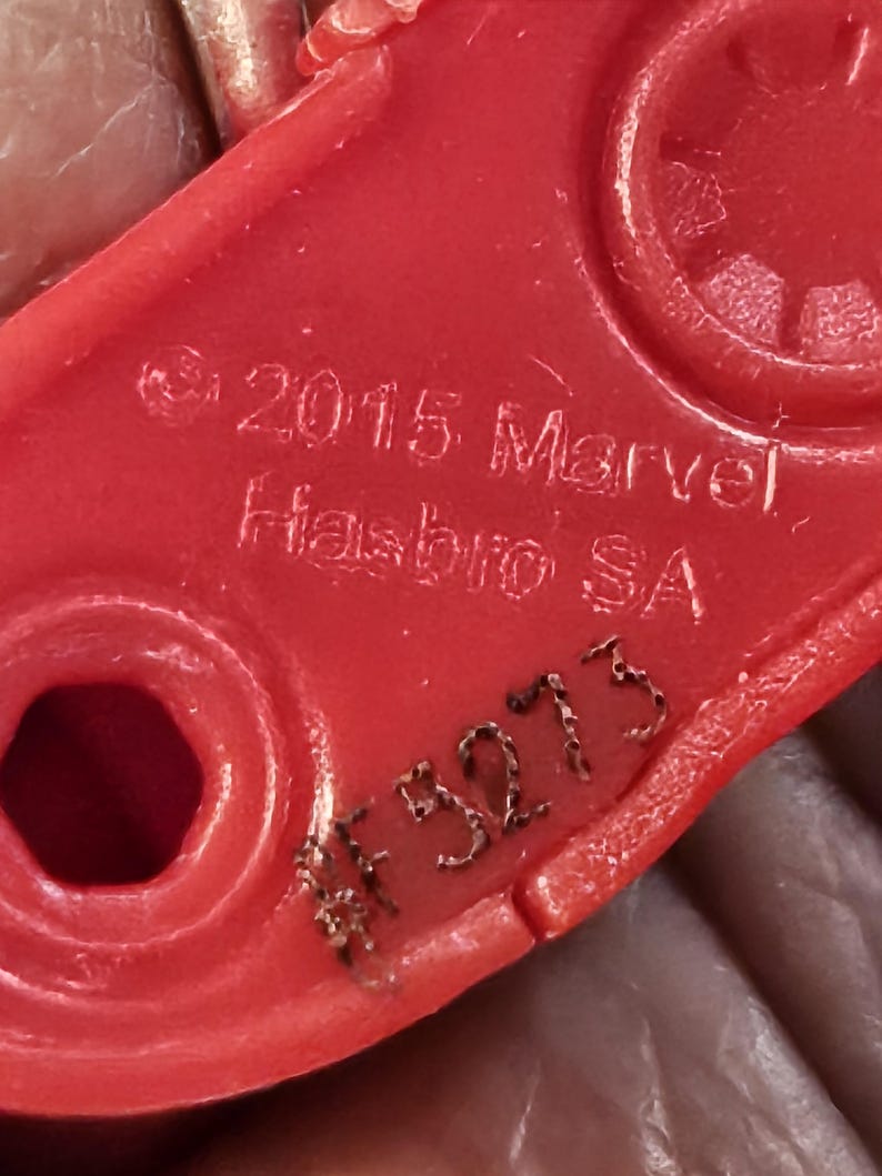 Może przedstawiać: Zbliżenie czerwonej plastikowej części zabawki z napisem "&copy; 2015 Marvel Hasbro SA" i numerem "#F3273" wygrawerowanym na powierzchni. Część ma zaokrąglony prostokątny kształt z okrągłymi i sześciokątnymi otworami.