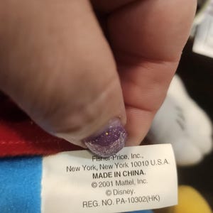 May include: A close-up of a white tag with black text from a Fisher-Price toy. The tag includes the company's address, the phrase "MADE IN CHINA," and copyright information for Mattel and Disney. The tag is held by a hand.